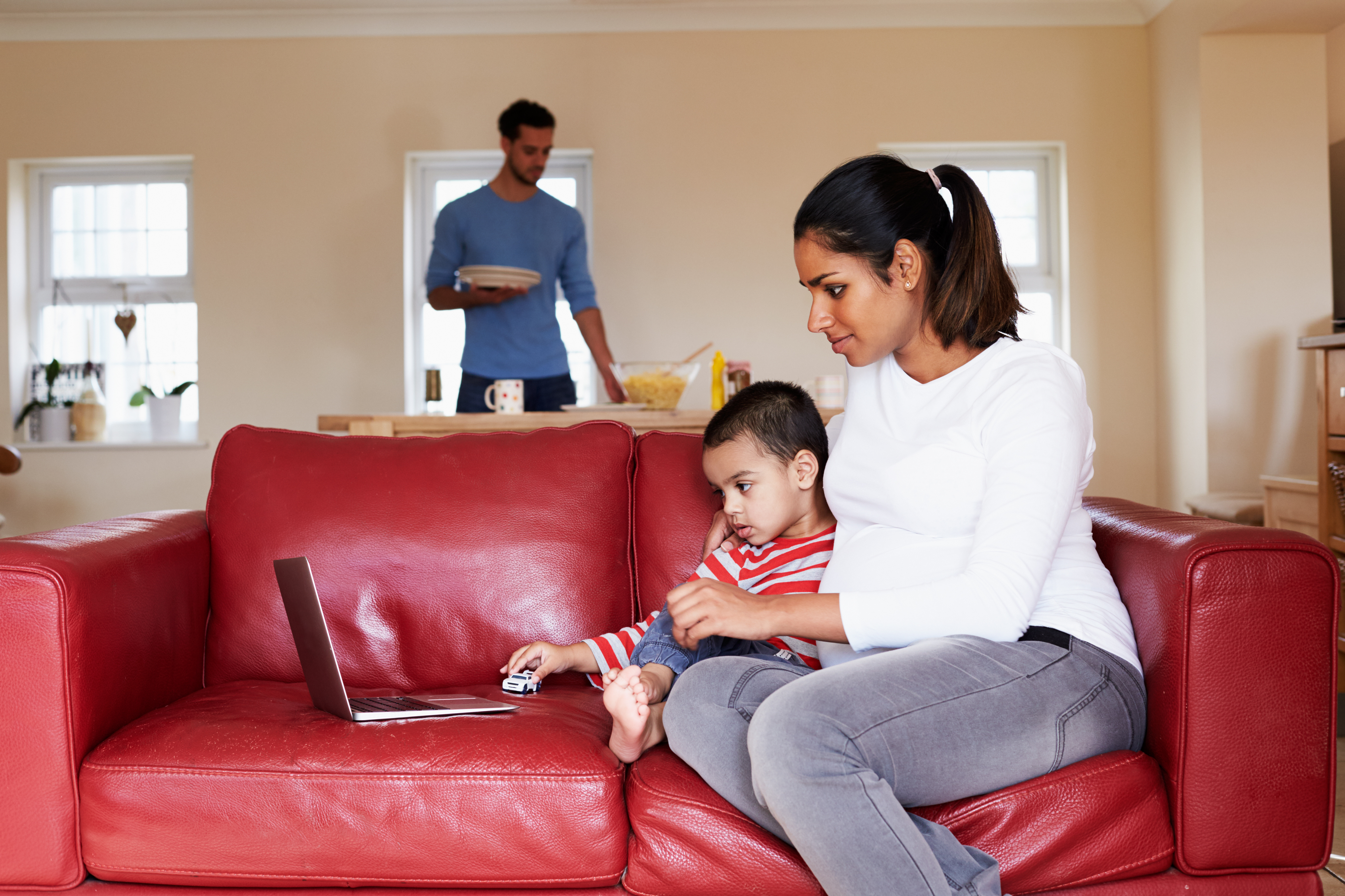 Pregnant Mother Looks At Laptop On Sofa With Son